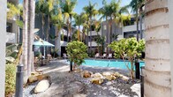 This property features a well-appointed outdoor courtyard centered around a generously sized, kidney-shaped swimming pool with clear turquoise water. The pool deck is finished in stamped concrete with a natural stone pattern, lending a sophisticated yet relaxed aesthetic. Surrounding the pool, an abundance of tall palm trees and tropical plantings, including banana leaf plants and dense shrubs, create a lush, resort-like atmosphere. Large natural boulders are incorporated into the landscaping beds, adding organic texture throughout the space.

The poolside area is furnished with slatted teak-style lounge chairs accented with striped cushions, complemented by teal market umbrellas that provide shade. Additional seating is arranged around the perimeter, offering multiple areas to relax. The courtyard is enclosed by a black iron fence and is embraced by the multi-story building façade, finished in cool gray tones, which provides a sense of privacy.

Access to the pool area is through a modern, glass-enclosed lobby corridor with polished concrete-style flooring, recessed lighting, and contemporary gold accent décor. The clean, minimalist interior design of the entryway contrasts elegantly with the vibrant outdoor greenery visible beyond.