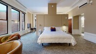 Located at 888 Delaware Ave in Buffalo, NY, this thoughtfully designed space blends historic architectural character with modern comfort across two bedrooms and one bathroom. The generously proportioned primary bedroom serves as the centerpiece of the layout, featuring an elegant panel-style headboard wall, warm neutral tones, plush carpeting, and crown molding that speaks to the building's storied heritage. Flanking wall sconces provide soft, ambient lighting, while a dedicated workspace with a desk and chair adds functional versatility. A cozy seating area with leather accent chairs and a decorative marble fireplace enhances the room's refined atmosphere. Natural light filters in through tall windows, offering views of the surrounding streetscape. A wall-mounted flat-screen television and a wall-mounted climate control unit ensure modern convenience. The second bedroom continues the warm, neutral aesthetic with carpeted flooring, crown molding, detailed wall paneling, and its own fireplace — creating an inviting retreat. The bathroom is finished with marble-tile flooring and features a full glass-enclosed walk-in shower, offering a clean, spa-like feel. Throughout the space, consistent warm beige tones, classic millwork, and rich wooden doors unify the design into a cohesive and comfortable environment.