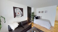 This single-floor studio presents an open-concept layout that thoughtfully combines living and sleeping areas within one cohesive space. The interior is immediately distinguished by a striking large-scale black-and-white pop-art portrait with a bold red accent, commanding attention on the main wall and setting an artistic, contemporary tone throughout.

The living area features a dark leather sofa paired with a patterned area rug, creating a grounded and comfortable lounge zone. Warm parquet flooring runs throughout, lending the space a classic yet inviting character. A dark decorative room divider subtly defines the transition between the living and sleeping areas, adding architectural interest without fully enclosing the space.

The sleeping area is furnished with a white daybed accented with black, grey, and patterned throw pillows, offering a clean and restful retreat. A tall indoor plant introduces a natural, organic element that softens the predominantly monochromatic palette. Two framed art pieces displayed on the adjacent wall contribute to the gallery-like aesthetic carried throughout the space.

The textured ceiling and white walls provide a neutral backdrop that allows the carefully curated furnishings and artwork to take center stage, resulting in a space that feels both expressive and functional.