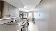 Located at 1150-1160 Rue Sherbrooke Est in Montréal, this shared laundry room is a well-equipped, single-floor facility designed for efficient, communal use. The room is outfitted with multiple front-loading LG washing machines positioned along one wall, complemented by stacked washer-dryer units that maximize the use of vertical space. Each machine features a digital control panel, offering modern convenience to users. A utility sink is also present, providing additional functionality for laundry needs. Flat, stainless steel folding surfaces sit atop the machines, offering practical workspace for sorting and folding laundry. A freestanding stainless steel utility table near the window provides additional workspace. Informational and instructional posters are mounted on the walls, offering guidance on machine operation and laundry best practices. The room is finished with light-coloured ceramic floor tiles that are easy to maintain and keep clean. Natural light filters in through a window that also offers a view of the surrounding neighbourhood. Overhead flush-mount lighting ensures the space is well-lit at all times. The neutral-toned walls and clean layout contribute to a functional, no-frills environment that prioritizes practicality and ease of use.