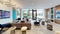 Located at 301 M Street in Washington, DC, this single-floor amenity space offers a thoughtfully designed environment that blends functionality with sophisticated contemporary style. Floor-to-ceiling windows flood the interior with natural light and provide views of lush greenery and a landscaped courtyard, creating a seamless connection between indoors and outdoors.

The co-working area features a sleek, dark communal table equipped with desktop computers and task lighting, set against a backdrop of warm wood-paneled walls and a vibrant large-scale abstract painting. A white open shelving unit adds an architectural element while displaying curated décor.

The lounge area offers generous sectional sofas in neutral tones, complemented by patterned accent stools and mixed metal side tables. Built-in wood and white shelving units with teal accent backing display art and books, adding depth and character to the space.

A media lounge features a wall-mounted flat-screen TV flanked by teal vertical panels and warm wood accents, illuminated by a striking multi-arm chandelier overhead.

The kitchenette and bar area includes a wood-and-white counter with bar seating, dual mounted televisions, and a full service counter with cabinetry — ideal for casual gatherings. Throughout the space, large-format tile flooring, recessed lighting, and a cohesive teal, walnut, and neutral palette create a polished and inviting ambiance.