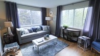 This single-floor space offers a thoughtfully designed interior that balances comfort and contemporary style. The living area serves as the focal point of the space, immediately drawing attention with its generous natural light streaming in through multiple large windows dressed in elegant navy blue geyelet curtains. The windows frame lush greenery outside, creating a seamless connection between the interior and the natural surroundings.

The living room features warm hardwood flooring that anchors the space with a sense of warmth and durability. A plush, textured grey area rug defines the seating area, complementing the light grey sofa adorned with an array of decorative blue and botanical-patterned throw pillows. A sleek white coffee table with chrome accents sits at the center, adding a modern touch.

Decorative wall art in a sunburst motif adds visual interest above the sofa, while a classic floor lamp provides soft ambient lighting. A dark wood console table positioned near the window offers additional display space, currently home to potted greenery that enhances the fresh, inviting atmosphere. The overall ambiance of the space is calm, curated, and filled with natural light, making it a welcoming environment throughout the day.