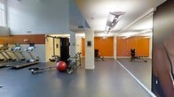 This fitness center is a thoughtfully designed workout space that combines functionality with an energizing atmosphere. The room is defined by bold terracotta-orange accent walls that create a motivating environment, contrasted against neutral gray and white surfaces throughout. Large floor-to-ceiling mirrors line one wall, visually expanding the space and allowing exercisers to monitor their form during workouts. The flooring is a durable, dark rubberized surface well-suited for gym use. The space is equipped with a multi-function cable strength training machine, a stability ball, and a dumbbell rack with a weight bench, providing a versatile range of exercise options for both strength and functional training. Recessed and panel lighting illuminates the area evenly from above, ensuring excellent visibility throughout. Structural columns are integrated into the layout, adding an architectural character to the open floor plan. A dedicated area near the mirrors offers generous open space, ideal for stretching, bodyweight exercises, or cardio activities. The high ceiling height contributes to an airy, open feel, making the space comfortable and inviting for daily fitness routines.