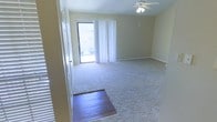 This single-floor space offers a clean, functional layout designed for comfortable everyday living. The main living area is the focal point of the home, characterized by neutral, warm-toned walls that create a welcoming and versatile atmosphere. Plush wall-to-wall carpeting covers the majority of the living area, providing a soft underfoot feel, while a section of rich hardwood flooring near the entryway creates a natural transition between spaces. A ceiling fan with an integrated light fixture is centrally mounted overhead, ensuring year-round comfort and ambient lighting throughout the room. Natural light pours in through a large sliding glass door dressed with sheer white curtains, which opens to an outdoor balcony area, offering views of the surrounding neighborhood. The sliding door serves as both a practical access point and a bright architectural feature that enhances the open feel of the living space. Louvered closet doors are visible along one wall, providing ample built-in storage. The space features clean baseboards and a smooth ceiling with a subtle textured finish, contributing to a tidy and well-maintained appearance throughout. Overall, the space presents a bright, airy, and move-in ready environment.