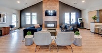 This contemporary single-floor clubhouse is designed to accommodate a variety of social and recreational needs. The centerpiece of the space is a sweeping vaulted lounge area anchored by soaring cathedral ceilings with exposed dark wood beams, creating a striking architectural focal point. A floor-to-ceiling wood-clad accent wall frames a linear fireplace and a wall-mounted television, offering a warm and inviting gathering spot. The lounge is furnished with an eclectic mix of seating, including leather sofas, plush armchairs, and ottomans arranged for easy conversation.

Adjacent to the lounge, an open-concept kitchen features white upper cabinetry contrasted by dark lower cabinets, stainless steel appliances, subway tile backsplash, and a large peninsula with pendant lighting and bar stool seating. A paw print jar on the counter hints at pet-friendly amenities nearby, including a dedicated pet prep area with dark cabinetry, stone countertops, and a sage green accent wall.

The open flex area offers additional seating arrangements alongside high-top bistro tables, ideal for casual work or socializing. A standout feature is the private media room, accessed through dramatic sliding dark barn doors, which reveals a cozy lounge with a wall-mounted screen. Light wood-look flooring flows consistently throughout, tying the entire space together with a clean, modern aesthetic.