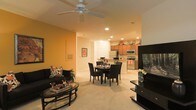 This single-floor residence offers a well-designed open-concept layout that connects the living, dining, and kitchen areas into one cohesive, flowing space. The living area features plush carpeting, warm amber-toned walls, and a ceiling fan with recessed lighting overhead, creating a comfortable and inviting atmosphere. A sleek entertainment setup anchors one side of the room, complemented by a stylish sofa and a glass-top coffee table. The dining area sits centrally within the open floor plan, featuring a dark wood dining table with matching chairs, ideal for hosting and everyday meals. Beyond the dining space, the kitchen is well-appointed with stainless steel appliances, including a refrigerator, microwave, and range, set against warm wood cabinetry and recessed lighting. The kitchen includes a breakfast bar that further connects the cooking and dining spaces. Natural light filters in through the windows, enhancing the bright and airy feel of the home. The thoughtful layout, neutral flooring, and consistent warm color palette throughout create a harmonious and functional living environment suited for both relaxing and entertaining.