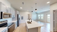 Located in Hutto, TX, this single-floor unit offers a thoughtfully designed open-concept layout that maximizes every square foot. Light hardwood-style flooring flows seamlessly throughout, complementing the bright, neutral color palette that enhances the sense of openness.

The kitchen is a standout feature, showcasing gray shaker-style cabinetry with black hardware, white quartz countertops, and a subway tile backsplash. A central island with a built-in sink and pendant lighting above serves as both a functional workspace and a casual gathering spot. Stainless steel appliances — including a refrigerator with ice maker, electric range, microwave, and dishwasher — complete the well-equipped kitchen.

The living area is bathed in natural light from large windows with views of the surrounding landscape, and a ceiling fan adds comfort to the space. A glass door opens directly to a private balcony, extending the living space outdoors.

The bathroom features a full tub-shower combo with clean white finishes and matte black fixtures and grab bars. A dedicated laundry room houses a full-size side-by-side washer and dryer with overhead shelving for additional storage. Recessed lighting and ceiling-mounted fixtures provide ample illumination throughout the unit.