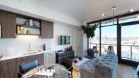 Located at 300 14th Street in San Diego, this single-floor residence offers a thoughtfully designed open-concept layout that seamlessly integrates the kitchen, dining, and living areas. The kitchen features dark wood cabinetry, white subway tile backsplash, stainless steel appliances, and a compact sink setup, providing a functional yet refined cooking space. A glass dining table with sleek chairs sits adjacent to the kitchen, creating an effortless transition into the living area. The living space is anchored by a tufted light grey sofa, a white stone coffee table, and a wall-mounted flat-screen television. A geometric-patterned credenza adds a contemporary design accent to the space. A large potted fiddle-leaf fig tree brings a touch of greenery, enhancing the modern aesthetic. Floor-to-ceiling sliding glass doors flood the interior with natural light and open to a private balcony overlooking the San Diego skyline, offering expansive city views. The herringbone-patterned hardwood flooring runs throughout, adding warmth and texture to the overall design. The space presents a cohesive blend of modern finishes and urban sophistication.