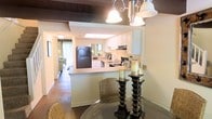 This two-story space offers a well-connected layout that blends the dining and kitchen areas into a cohesive, functional living environment. The dining area is anchored by a glass-top table and complemented by an elegant chandelier with bell-shaped shades, adding a classic touch to the space. Decorative candle stands on the table contribute to the warm, intimate ambiance.

The kitchen is designed with a pass-through counter that opens toward the dining area, promoting an open-concept feel. White cabinetry provides ample storage, while the countertops offer generous workspace. The kitchen is equipped with a full suite of appliances, including a large refrigerator, stove, and built-in microwave. A skylight above the kitchen allows natural light to filter in, brightening the workspace. Dark wood beam accents on the ceiling add a rustic architectural contrast to the otherwise light interior.

Hardwood flooring runs throughout the main living areas, adding warmth and continuity. Beyond the kitchen, a glimpse into the living area reveals natural light streaming in through sliding glass doors, suggesting an inviting and well-lit atmosphere throughout the home.