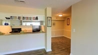 This single-floor residence offers a thoughtfully designed layout that balances functionality and comfort. The interior is anchored by rich, dark hardwood-style flooring that flows continuously throughout the main living areas, lending warmth and cohesion to the space.

A standout architectural feature is the built-in pass-through opening between the kitchen and the adjacent living area, complete with a granite-style countertop ledge and open shelving above — ideal for displaying books, décor, and everyday essentials. This design element not only enhances connectivity between spaces but also allows natural light from the kitchen window to filter into the main living area.

The walls are finished in a soft, neutral tone that creates a calm and inviting ambiance throughout. White baseboards and trim provide clean contrast and a classic finishing touch. The open passageway leading to the dining area features a textured ceiling and flush-mount ceiling light, creating a well-lit and welcoming transition between rooms.

Decorative wall art and a sunburst mirror accent the hallway, adding personality and visual interest. The dining area is open and spacious, offering ample room for a full dining set. Overall, the space presents a practical yet charming interior with classic detailing and a warm, livable atmosphere throughout.
