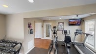 This fitness center is a purpose-built exercise space designed to provide residents or users with a convenient and functional workout environment. The room features a clean, neutral color palette with beige walls and white trim, creating a bright and motivating atmosphere enhanced by recessed ceiling lighting. The flooring transitions from hardwood near the entrance to durable dark rubber or gym-grade matting in the main exercise area, providing both practicality and comfort underfoot. The space is equipped with a selection of cardio machines, including an elliptical trainer and a recumbent exercise bike, positioned to allow for comfortable use. A wall-mounted flat-screen television is installed at an ideal viewing height, enabling users to stay entertained during workouts. Fitness-related informational posters are displayed on the walls, adding both function and visual interest. A water cooler station is conveniently mounted near the entrance, ensuring hydration is easily accessible. The space includes a mirrored door, adding depth and utility to the room. A clearly labeled "Fitness Center" sign is posted near the entrance, and the overall layout is organized and easy to navigate, making this a practical and well-appointed facility for everyday exercise.