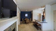 This single-floor residence offers a thoughtfully designed open-concept layout that seamlessly connects the living, dining, and kitchen areas. The living room serves as a central focal point, anchored by a fireplace with a classic white mantel and a slate tile surround in earthy tones. A wall-mounted flat-screen television is positioned above the mantel, and crown molding runs along the ceiling, adding a refined architectural detail. A ceiling fan contributes to the room's comfort and airflow.

The dining area is defined by a bold navy blue accent wall, which provides a striking contrast to the neutral, light-toned carpet and cream walls throughout the space. A round dining table surrounded by upholstered chairs sits comfortably within this area, with a small window allowing natural light in.

Adjacent to the dining space, the kitchen bar features dark countertops and is lined with leather-upholstered bar stools, offering casual seating. Pendant lights hang above the bar, providing warm, focused illumination. The kitchen area, partially visible, includes cabinetry and additional track lighting overhead. Overall, the space presents a cohesive and functional design with warm, neutral tones accented by rich, deep colors.