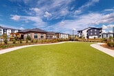 Located at 100 Oakbrook Drive in McDonough, Georgia, this thoughtfully designed outdoor amenity space serves as a central gathering area within a modern residential community. At its heart lies a generously sized putting green with a distinctive organic shape, surrounded by a smooth concrete pathway that winds gracefully throughout the grounds. The lush, well-maintained turf creates a vibrant focal point visible from multiple vantage points across the property.

Meticulously curated landscaping frames the space, incorporating ornamental grasses, low-lying shrubs, mulched garden beds, and young trees that will mature to provide additional shade and character over time. Black wrought-iron fencing defines the perimeter, lending a clean, classic aesthetic while maintaining open sightlines to the surrounding greenery and towering pine trees that form a natural backdrop to the north.

The single-story brick clubhouse anchors one side of the amenity area, featuring large windows and a covered porch that overlooks the green. The broader community context is visible throughout, with multi-story residential buildings providing an architecturally cohesive backdrop. Wide-open skies above and abundant natural light enhance the overall ambiance, making this outdoor space an inviting and functional retreat for community residents year-round.