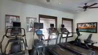 This fitness center is a dedicated, single-floor exercise space designed to support a range of cardio-focused workouts. The room is outfitted with multiple treadmills and elliptical trainers, each equipped with individual digital display screens, allowing users to track their workout metrics or enjoy entertainment while exercising. Natural light filters through several wood-framed windows dressed with horizontal blinds, lending the space a bright and welcoming atmosphere. The walls are finished in a clean, neutral tone, contributing to an open and airy feel. A ceiling fan overhead aids in air circulation, helping to keep the environment comfortable during workouts. An informational fitness chart is mounted on the wall near the entrance, providing exercise reference and guidance for users. The flooring is dark-toned, offering a practical and durable surface suited for high-traffic gym use. The layout is thoughtfully arranged to allow ease of movement between machines. Overall, this fitness center presents a functional and well-maintained environment, ideal for residents or guests seeking a convenient space to maintain their health and wellness routines.