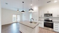 Located at Harbor Pointe Cir in Fruitland Park, FL, this single-story, 2-bedroom, 2-bathroom home offers a clean, modern design with thoughtful finishes throughout. The open-concept layout seamlessly connects the living room, dining area, and kitchen, all finished with warm wood-look flooring and bathed in natural light from French doors that lead to a private patio. The kitchen is a standout feature, boasting a large center island with a quartz countertop and undermount sink with a matte black faucet, white shaker cabinetry, subway tile backsplash, stainless steel appliances, and striking industrial-style pendant lighting. Both bedrooms are carpeted and include ceiling fans, with the primary bedroom featuring a spacious walk-in closet with built-in wire shelving. Each bathroom is finished with quartz countertops, white shaker vanities, undermount sinks, matte black fixtures, and large mirrors with vanity lighting. A dedicated in-unit laundry room houses a full-size washer and dryer. Additional conveniences include a pantry, utility room, and hallway connecting all spaces efficiently. The home's bright white walls, black hardware accents, and consistent finishes create a cohesive, contemporary aesthetic throughout.