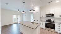 Located at Harbor Pointe Cir in Fruitland Park, FL, this single-story, 2-bedroom, 2-bathroom home offers a clean, modern design with thoughtful finishes throughout. The open-concept layout seamlessly connects the living room, dining area, and kitchen, all finished with warm wood-look flooring and bathed in natural light from French doors that lead to a private patio. The kitchen is a standout feature, boasting a large center island with a quartz countertop and undermount sink with a matte black faucet, white shaker cabinetry, subway tile backsplash, stainless steel appliances, and striking industrial-style pendant lighting. Both bedrooms are carpeted and include ceiling fans, with the primary bedroom featuring a spacious walk-in closet with built-in wire shelving. Each bathroom is finished with quartz countertops, white shaker vanities, undermount sinks, matte black fixtures, and large mirrors with vanity lighting. A dedicated in-unit laundry room houses a full-size washer and dryer. Additional conveniences include a pantry, utility room, and hallway connecting all spaces efficiently. The home's bright white walls, black hardware accents, and consistent finishes create a cohesive, contemporary aesthetic throughout.