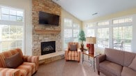 This single-floor home offers a thoughtfully designed layout that balances comfort and functionality. The living area is anchored by a dramatic floor-to-ceiling stacked stone fireplace, complete with a wood mantel and wall-mounted television, set beneath soaring vaulted ceilings. A ceiling fan enhances airflow throughout the open space, which features plush carpeting and an abundance of natural light streaming through multiple windows. Adjacent to the main living area, a sun-drenched sitting room is enclosed by expansive windows on multiple walls, offering sweeping outdoor views of lush greenery. This bright retreat provides a tranquil space ideal for relaxation. The open-concept layout flows seamlessly into a well-appointed kitchen, featuring warm honey-toned wood cabinetry, generous counter space, tile flooring, and a full suite of stainless steel appliances including a double oven, built-in microwave, dishwasher, and French door refrigerator. A dedicated dining area with tile flooring and large windows overlooking the outdoors completes the kitchen zone. Throughout the home, soft yellow walls, white trim, and recessed lighting contribute to a bright, welcoming ambiance that feels both homey and inviting.