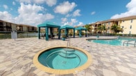 This expansive outdoor amenity space is designed to deliver a resort-like experience within a residential community setting. The centerpiece is a large, free-form swimming pool with clear turquoise water, surrounded by a generously sized paver deck finished in a multi-toned stone pattern that adds visual texture and sophistication underfoot. Adjacent to the pool sits a circular hot tub, elegantly inset into the pavement and accessible via stainless steel handrails.

A row of freestanding teal cabanas with coordinating drape curtains lines one side of the pool deck, each furnished with seating and shaded lounge areas ideal for relaxation. Additional chaise lounges are arranged throughout the deck, offering multiple vantage points overlooking the water. Lush tropical landscaping, including tall palm trees, colorful flowering shrubs, and manicured plant beds, frames the space and reinforces its Florida-inspired aesthetic.

A community building with floor-to-ceiling glass doors and large windows borders the deck, providing direct pool access while flooding the interior with natural light and views of the outdoor space. A gated perimeter opens toward a waterfront greenway, adding an additional layer of scenic backdrop to the overall ambiance.