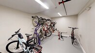 This 150 square foot room is a thoughtfully designed bicycle storage area, maximizing space efficiency in a compact footprint. The room features bright, even lighting provided by ceiling-mounted fluorescent fixtures, illuminating the entire space effectively. The walls are painted in a light, neutral color, creating a clean and spacious feel.

The most prominent feature is the wall-mounted bicycle storage system. Multiple bikes are securely hung vertically along one wall, utilizing vertical space efficiently. This rack system accommodates various bicycle types and sizes, from road bikes to mountain bikes. The opposite wall features additional horizontal racks for more storage options.

The floor is finished with a smooth, durable material, likely epoxy or similar, providing easy cleaning and maintenance. This surface also allows for effortless movement of bicycles in and out of the space. There's ample floor area for bicycle maintenance or additional storage of cycling gear.

The room's layout is well-organized, with clear pathways for accessing stored bikes. A ceiling-mounted fire sprinkler system is visible, ensuring safety compliance. The space also includes a wall-mounted smoke detector for added security.

This bicycle storage room demonstrates efficient use of limited square footage, providing a practical and organized solution for multi-bike storage in a residential or commercial setting.