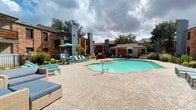 This outdoor amenity area centers around a generously sized, freeform swimming pool with clear turquoise water, framed by a wide concrete sundeck designed for relaxation and social gatherings. The pool's geometric shape accommodates both leisure swimming and lounging, with clearly marked depth indicators along the tiled coping edge. Surrounding the pool, an array of cushioned chaise lounges and wicker seating arrangements are accented by teal umbrellas, providing shaded retreats throughout the day.

The complex features classic brick construction with bold gray chimney towers that serve as distinctive architectural landmarks. A dedicated pool house structure sits adjacent to the pool area, offering additional covered seating and direct access to the water. Mature trees and well-maintained landscaping line the perimeter, adding privacy and greenery to the outdoor environment.

Pool access is managed through a secure entry vestibule equipped with a glass-block sidelight panel, a glass sliding door, security camera, and an emergency 911 telephone station, underscoring the safety infrastructure of the space. The overall ambiance balances a resort-like aesthetic with practical, community-oriented amenities.
