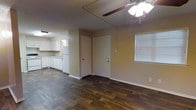This single-floor space offers a straightforward, functional layout with an open living area that flows directly into the kitchen. The living area features rich, dark hardwood-style flooring that contrasts with the neutral beige walls, creating a warm and grounded atmosphere. A ceiling fan with a light fixture is centrally mounted, providing both air circulation and overhead lighting. A window fitted with white horizontal blinds allows natural light to filter into the space.

The kitchen, visible from the living area, is outfitted with white cabinetry that extends along the walls, offering ample storage. White appliances, including a refrigerator and additional countertop units, complement the clean, cohesive look of the kitchen. The transition between the two areas is seamless, making the space feel connected and open.

A white paneled door leads to an additional room or exterior access point, adding functionality to the layout. White baseboards throughout provide a clean finishing detail. The textured ceiling adds a subtle architectural element. Overall, the space presents a clean, move-in-ready environment with practical features suited for everyday living.