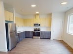 This 990-square-foot, two-story home at 1047 Tartarian Ave in Richland, WA offers a well-organized layout across two floors. The ground floor (350 sq ft) features a combined kitchen and living room, providing an open, functional living area. The kitchen is outfitted with stainless steel appliances including a refrigerator, range, microwave, and dishwasher, complemented by two-tone cabinetry — light wood upper cabinets and dark wood-grain lower cabinets — set against clean white countertops. Wood-look flooring runs throughout the home, adding warmth and consistency. The upper floor (640 sq ft) contains both bedrooms, two full bathrooms, a hallway, and a staircase. The primary bedroom spans 130 square feet and, like the second bedroom, features neutral white walls, generous natural light from large windows with outdoor views, and sliding double-panel closet doors. Both bathrooms are finished with matching wood-grain vanities, white tub/shower combos, and light flooring. The home's clean, modern aesthetic is defined by its neutral color palette, recessed lighting, and consistent use of materials, making it a bright, move-in-ready residence.