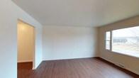 This two-floor space offers a straightforward and functional layout with a clean, neutral aesthetic throughout. The main living area is generously proportioned, featuring warm-toned wood-style flooring that runs continuously across the room, providing both durability and visual warmth. Crisp white walls and white trim create a bright, airy atmosphere, while a window allows natural light to filter into the space, enhancing its open feel. The room's simple design provides a versatile canvas adaptable to a variety of furnishing styles. An open doorway connects to an adjacent area, suggesting a practical flow between spaces. The clean baseboards and smooth ceiling contribute to the overall polished, well-maintained appearance of the interior. Spanning two floors, the property offers separation between living and private spaces, a layout that supports both daily functionality and privacy. The consistent use of neutral tones and hard-surface flooring throughout gives the space a cohesive, move-in-ready quality suitable for a range of occupants.