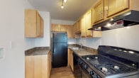 This two-floor residence offers a thoughtfully designed layout that balances functionality and comfort. The kitchen is efficiently arranged with warm honey-toned wood cabinetry extending to the ceiling, providing generous storage space. Dark granite-style countertops complement the full suite of black appliances, including a gas range, refrigerator, and built-in range hood. A stainless steel sink is positioned beneath a window, and track lighting illuminates the workspace. Rich hardwood flooring runs throughout the kitchen and continues into the main living areas, adding warmth and visual continuity to the home. The open-plan living and dining area is bright and airy, with crisp white walls and a neutral palette that create a clean, welcoming atmosphere. A pass-through opening connects the spaces seamlessly, while a glass-paneled exterior door at the far end allows natural light to flood in, brightening the interior. An elegant chandelier adds a refined touch to the dining area. HVAC vents are integrated into the floor and walls, ensuring year-round comfort. The overall design of this two-floor home reflects a practical yet inviting character, with classic finishes and well-proportioned spaces suited for everyday living.