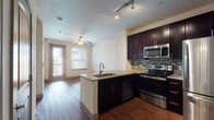 This single-floor residence is designed with a modern aesthetic and functional layout. The open-concept kitchen serves as a central feature, showcasing rich dark cabinetry extending to the ceiling, granite countertops, and a full suite of stainless steel appliances including a range, microwave, and refrigerator. A mosaic tile backsplash adds visual interest, while a peninsula with a granite surface and built-in sink provides additional workspace and separates the kitchen from the living area. Track lighting and hardwood flooring carry throughout the main living space, which benefits from natural light streaming through windows and a glass door leading to a balcony. A ceiling fan keeps the living area comfortable. A hallway connects to the remaining rooms, including a carpeted bedroom featuring dual windows with city views and its own ceiling fan. The bathroom is well-appointed with dark cabinetry, a granite-topped vanity, a framed mirror with overhead lighting, and tile flooring. A dedicated laundry area with washer and dryer hookups adds convenience. The overall ambiance is contemporary and cohesive, with consistent finishes throughout.