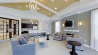 This single-story space showcases an open-concept design that seamlessly connects the living, dining, and kitchen areas into one cohesive environment. The living area is anchored by a classic fireplace with a shiplap-style surround, complemented by a wall-mounted flat-screen television and a pair of upholstered accent chairs. A large sectional sofa in soft gray tones defines the seating area, centered around a sleek glass coffee table. A geometric chandelier serves as a striking focal point overhead, drawing the eye upward to the soaring vaulted ceiling, which features a raised tray section finished in warm gold tones and lined with recessed lighting. The kitchen is outfitted with dark cabinetry, stainless steel appliances, and a white countertop breakfast bar with bar stool seating. Pendant lights hang above the bar, adding warmth to the space. Neutral-toned carpeting flows throughout the living area, while soft gray walls and white trim provide a clean, contemporary aesthetic. Natural light filters in through windows dressed with white curtains, contributing to the bright and welcoming ambiance of the space.