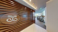 This sophisticated fitness center is defined by its modern architectural design and exceptional range of amenities. The entry makes an immediate impression with a boldly slatted wood-paneled accent wall bearing a branded logo, setting an upscale tone throughout. Floor-to-ceiling windows line the perimeter, flooding the space with natural light and offering lush views of tropical palm trees and surrounding greenery.

The main workout floor is generously proportioned, accommodating a comprehensive array of Cybex strength machines, cable systems, and free weights including a fully stocked dumbbell rack. A dedicated functional training zone features multi-station rigs, a heavy punching bag, TRX suspension systems, and barbells alongside a polished marble accent wall. Cardio equipment, including treadmills, ellipticals, and stair climbers, is positioned along the window-facing walls to maximize outdoor views during workouts.

A relaxation and recovery lounge includes warm wood cabinetry, a beverage refrigerator, high-top seating, and a vibrant living green wall. Glass doors reveal an adjacent indoor pool area, elegantly enclosed with a sparkling overhead installation. Recessed and ambient lighting throughout enhances the clean, white-and-neutral palette, creating a bright and motivating environment.