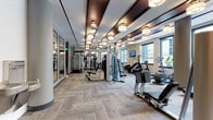 Located at 1900 Chapman Avenue in Rockville, MD, this well-appointed fitness center offers a comprehensive workout environment designed with both function and style in mind. The main exercise floor is generously proportioned, featuring a sophisticated ceiling treatment of dark wood beam accents complemented by rows of elegant drum pendant light fixtures that cast a warm, inviting glow throughout the space. Large windows line the perimeter, flooding the interior with natural light and providing views of the surrounding outdoor areas.

The main floor is equipped with an extensive array of cardio machines, including treadmills, ellipticals, stair climbers, and stationary bikes, all positioned to take advantage of the natural light. A dedicated strength training zone houses cable machines, a Smith machine, free weights, medicine balls, and a variety of functional training equipment. Full-length mirrors are strategically placed throughout, enhancing the sense of space and supporting proper form during workouts.

A separate spin studio features light hardwood flooring, a striking geometric ceiling grid adorned with globe pendant lights, wall-to-wall mirrors, and multiple stationary cycling bikes — creating an energetic and focused environment for cycling classes. Additional amenities include water fountains, personal storage cubbies, and wall-mounted televisions. The neutral-toned carpet, modern color palette, and refined architectural details give the entire facility a polished, upscale character.