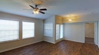 This single-floor space presents a practical and well-organized layout spread across several interconnected areas. The main living area is a notable highlight, featuring rich, dark wood-style flooring that runs throughout much of the unit. Multiple windows fitted with white horizontal blinds allow for generous natural light, creating a bright and open atmosphere. A ceiling fan with an integrated light fixture is centrally positioned in the living area, complemented by a pendant light that adds warmth to the space.

The kitchen area is equipped with an island or central work surface, providing additional prep and workspace. Cabinetry lines the walls, offering practical storage solutions. Adjacent to the kitchen, a compact bathroom is outfitted with a toilet and sink, efficiently utilizing the available space.

The floor plan, as seen in the overhead view, reveals a thoughtfully arranged layout with clearly defined zones for living, cooking, and bathing. The warm-toned walls pair with the wood-style flooring to give the space a cohesive and inviting character. Overall, the unit balances functionality with comfort in a compact, single-level footprint.