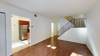 This two-story space offers a well-organized layout with distinct living areas spread across both floors. The main level features rich, dark hardwood flooring that runs throughout the open living area, reflecting natural light and adding warmth to the interior. Bright white walls create a clean, neutral backdrop that enhances the sense of openness throughout the space.

A decorative wrought iron staircase with ornamental scrollwork detailing serves as a standout architectural feature, connecting the main level to the upper floor. The staircase is positioned toward the rear of the living area, allowing the main space to remain unobstructed and functional.

Adjacent to the living area, the kitchen is partially visible and features warm wood cabinetry paired with stainless steel appliances, including a full-size refrigerator. The kitchen area has tile flooring, providing a practical contrast to the hardwood in the living space. A doorway near the staircase leads to an additional room, likely a bathroom or utility space, contributing to the overall functionality of the main level. Ceiling vents suggest a central HVAC system throughout the home.