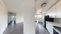 This single-floor unit offers a thoughtfully designed layout with a clean, contemporary aesthetic throughout. Dark laminate flooring contrasts beautifully with crisp white walls and trim, creating a modern and cohesive look across all rooms.

The kitchen is a standout feature, equipped with full-height white shaker-style cabinetry, a classic subway tile backsplash, and a full suite of stainless steel appliances including a refrigerator, electric range, and over-the-range microwave. A peninsula with an integrated sink and light countertop provides additional prep space and connects seamlessly to the open living area.

The spacious living room benefits from generous natural light, with large sliding doors and a secondary window offering views to an outdoor balcony area. A baseboard heating unit runs along the exterior wall, ensuring year-round comfort.

Two bedrooms are present, each finished with the same laminate flooring and neutral white walls. Both rooms are equipped with closets — one featuring a sliding door system and the other a paneled sliding door with a brushed metal handle — providing ample storage.

The bathroom is neatly appointed with large-format grey wall tiles surrounding a full soaking tub and shower combination, complemented by chrome fixtures and a window that brings in natural light.