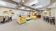 Located at 3021 Maple Valley Dr in Madison, Wisconsin, this single-floor community clubhouse offers a generously sized, open gathering space designed for socializing and recreation. The layout flows seamlessly from one area to the next, creating a cohesive and welcoming environment throughout.

The main lounge area is furnished with comfortable yellow upholstered sofas and chairs arranged around a soft green area rug, providing an inviting setting for relaxation and conversation. A large wall-mounted flat-screen TV anchors one side of the room, while large glass doors at the far end offer views of an outdoor pool, filling the space with natural light.

Adjacent to the lounge is a recreation zone centered around a full-sized pool table with a rich red felt surface, complemented by a cue rack and a bistro-style high-top table with bar stools — ideal for casual gatherings. A decorative fireplace adds a warm, homey touch to this area.

The kitchen is well-equipped with white cabinetry, a stainless steel refrigerator, and a range, set against sage green accent walls. A curved breakfast bar with padded swivel stools makes it perfect for entertaining. A dropped, coffered ceiling with recessed lighting and skylights runs throughout the space, adding architectural interest while keeping the atmosphere bright and airy.