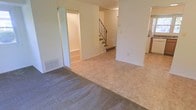 This two-story space presents a practical and open layout across its main level, with a staircase providing access to the upper floor. The main living area features a combination of carpet and tile flooring, creating distinct zones within the open floor plan. Neutral-toned walls throughout contribute to a clean, understated aesthetic. A doorway visible from the main living area leads to an adjacent room, adding to the functional flow of the space. The staircase, framed by a metal railing, serves as a central architectural element connecting the two floors. A floor-level air vent indicates a built-in HVAC system for climate control. Natural light filters into the space, enhancing the bright and airy feel of the interior. A glimpse of what appears to be a kitchen area is visible to the right, suggesting convenient adjacency to the living space. Overall, the layout is straightforward and versatile, lending itself well to a variety of uses across both floors.