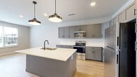 This well-appointed one-bedroom, one-bathroom unit offers a thoughtfully designed layout across a single floor, combining modern finishes with functional living spaces. The open-concept kitchen, dining, and living area serves as the heart of the home, featuring light hardwood flooring that flows seamlessly throughout. The kitchen is outfitted with gray shaker-style cabinetry, a white subway tile backsplash, stainless steel appliances including an electric range and built-in microwave, and a generous center island with a white quartz countertop, undermount sink, and matte black faucet. Stylish pendant lights hang above the island, adding a contemporary accent. The living area receives ample natural light through a large window with views of the surrounding neighborhood. The bedroom is a comfortable, well-sized retreat finished in soft neutral tones with natural light provided by a window overlooking the exterior. Two closets offer generous storage, including a walk-in closet equipped with wire shelving and hanging rods. The bathroom is a standout feature, showcasing a dual-sink vanity with gray cabinetry, white quartz countertops, matte black fixtures, and two backlit mirrors, complemented by a tall linen tower cabinet. A dedicated laundry area houses a stacked washer and dryer unit for added convenience.