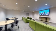 This single-floor communal space is thoughtfully designed to accommodate both relaxed social gatherings and productive group meetings. The layout is divided into two functional zones that flow seamlessly into one another, unified by a striking grey and yellow-green hexagonal patterned carpet that anchors the entire room.

The lounge area is defined by multiple modular sectional sofas upholstered in vivid olive green velvet, arranged to encourage conversation and comfortable viewing. Complementing the seating are sleek, low-profile black side tables. A large wall-mounted display screen serves as a focal point, supported by a slim console table, making this zone well-suited for presentations or casual entertainment. Abstract black-and-white artwork adorns the white walls, adding a curated artistic element to the space.

The meeting and dining zone features two round tables with light wood tops, each surrounded by modern black chairs, offering a practical setting for collaborative work or casual dining. Recessed ceiling lighting throughout provides consistent, even illumination across both areas, while in-ceiling speakers suggest integrated audio capability. The neutral white walls and clean architectural lines contribute to a bright, contemporary ambiance suitable for a shared amenity or collaborative workspace.