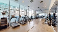 Located at 705 W 9th St in the heart of downtown Los Angeles, this modern fitness center offers a comprehensive workout facility within a thoughtfully designed single-level space. The gym's most striking architectural feature is its expansive floor-to-ceiling glass curtain wall, which floods the interior with abundant natural light while framing impressive views of the surrounding Los Angeles skyline.

The space is organized to accommodate a wide range of fitness activities. Along the windowed perimeter, a row of cardio machines — including treadmills, ellipticals, and stationary and rowing bikes — allows users to exercise while taking in the urban panorama. The strength training area is equally well-appointed, featuring a multi-station cable machine, a squat rack with an adjustable bench, and a dedicated free weights section stocked with dumbbells, medicine balls, and battle ropes.

Full-length mirrors line one wall, enhancing the sense of openness and allowing users to monitor their form. Recessed ceiling lighting supplements the natural light, ensuring a bright and energizing atmosphere throughout. The durable, easy-to-maintain flooring is consistent throughout the space, complementing the clean, contemporary aesthetic. The fitness center is accessed through glass-panel doors, reinforcing the sleek, modern design that defines the entire facility.