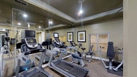 This fitness center offers a well-appointed space designed to support a variety of cardio-focused workouts. The room is furnished with multiple treadmills, upright stationary bikes, a recumbent bike, and an elliptical-style machine, providing residents with a comprehensive selection of equipment. Each cardio machine is equipped with an individual monitor, allowing users to track their workout metrics or enjoy entertainment while exercising. A large wall-mounted flat-screen television is also present, adding to the engaging atmosphere.

The interior design features warm olive-toned walls accented by classic crown molding and a tray ceiling, lending the space a polished, residential feel rather than a stark, utilitarian aesthetic. Large wall mirrors visually expand the room and allow users to monitor their form during exercise. Framed botanical artwork adds a decorative touch to the walls. Recessed and track lighting illuminate the space effectively, while a ceiling fan contributes to air circulation and comfort. The flooring consists of low-pile carpet, providing a practical and cushioned surface throughout. Overall, the fitness center balances functionality with thoughtful design, creating an inviting environment for daily exercise.