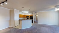 This single-floor residence offers a thoughtfully designed open-concept layout that seamlessly connects the kitchen and living areas. The kitchen is outfitted with warm honey-toned wood cabinetry, dark countertops, and black appliances including a range, built-in microwave, and refrigerator. A peninsula with pendant lighting above serves as both a functional workspace and a visual divider between the kitchen and the carpeted living room. Crown molding lines the ceilings throughout the main living area, lending a polished, refined character to the space. The living room is generously sized, anchored by a ceiling fan and bathed in natural light from a large sliding glass door that opens to a private balcony. The bedrooms are carpeted and feature vertical blinds over windows that allow for adjustable natural light. One bedroom includes a ceiling fan and access to a walk-in closet, while another offers direct access to a bathroom with warm wood vanity cabinetry. Additional storage is provided through well-proportioned closets. The tile flooring in the kitchen transitions cleanly to carpet in the living and sleeping areas, creating distinct functional zones throughout this comfortable and practical residence.