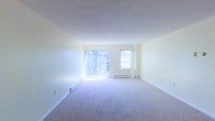 This single-floor space presents a spacious, open living area finished with wall-to-wall carpeting in a neutral tone, contributing to a clean and understated aesthetic. The room is bathed in natural light, primarily sourced from a large sliding glass door that opens to an outdoor area with visible greenery beyond, creating a pleasant connection to the outdoors. A secondary window positioned adjacent to the sliding door further enhances the brightness of the interior. The walls are painted in a soft, neutral hue, lending the space an airy and versatile feel that accommodates a variety of furnishing styles. Baseboard heating units are present along the walls, indicating a functional heating system integrated neatly into the room's perimeter. The ceilings are standard height with a smooth finish, maintaining a straightforward and practical design throughout. The overall layout is open and unobstructed, offering flexibility in how the space can be arranged and utilized. The combination of abundant natural light, neutral finishes, and outdoor views through the sliding glass door gives the space a welcoming and comfortable ambiance.