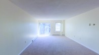 This single-floor space presents a spacious, open living area finished with wall-to-wall carpeting in a neutral tone, contributing to a clean and understated aesthetic. The room is bathed in natural light, primarily sourced from a large sliding glass door that opens to an outdoor area with visible greenery beyond, creating a pleasant connection to the outdoors. A secondary window positioned adjacent to the sliding door further enhances the brightness of the interior. The walls are painted in a soft, neutral hue, lending the space an airy and versatile feel that accommodates a variety of furnishing styles. Baseboard heating units are present along the walls, indicating a functional heating system integrated neatly into the room's perimeter. The ceilings are standard height with a smooth finish, maintaining a straightforward and practical design throughout. The overall layout is open and unobstructed, offering flexibility in how the space can be arranged and utilized. The combination of abundant natural light, neutral finishes, and outdoor views through the sliding glass door gives the space a welcoming and comfortable ambiance.