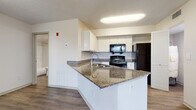 This single-floor residential unit offers a thoughtfully designed layout that balances functionality and comfort throughout its living spaces. The open-concept living area is finished with warm wood-look luxury vinyl flooring, neutral white walls, and a textured ceiling fitted with a ceiling fan, creating a bright and airy atmosphere. Natural light filters in through blinded windows, adding to the welcoming ambiance. The kitchen is a standout feature, showcasing granite countertops with a peninsular island that includes a built-in sink — ideal for both meal preparation and casual dining. White cabinetry provides ample storage, while black appliances, including a range, over-the-range microwave, and refrigerator, offer a clean contrast to the bright cabinetry and countertops. The kitchen flows seamlessly into the living space, enhancing the open feel of the unit. The bedroom is finished with plush carpet flooring and features a ceiling fan, a window with views of surrounding greenery, and generous closet storage with louvered bi-fold doors. The bathroom is clean and functional, equipped with a full soaking tub and shower combination, all finished in crisp white. Throughout the unit, consistent flooring, neutral tones, and practical design choices create a cohesive and move-in-ready living environment.