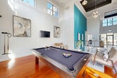 Located at 3378 Greenbriar Pkwy SW in Atlanta, GA, this thoughtfully designed community space makes a bold architectural statement from the moment you enter. The main gathering area commands attention with dramatic double-height ceilings, exposed wood beams, and an eye-catching teal accent wall adorned with framed artwork. Rich hardwood flooring flows throughout, complemented by abundant natural light pouring in through expansive clerestory and floor-level windows. A sleek cable-railing staircase with a wood-topped handrail anchors the central hallway, lending a contemporary edge to the space. The open living and dining area is generously proportioned, featuring a full-sized billiards table, modern pendant lighting, and a comfortable seating arrangement ideal for socializing. A bar-height dining table with white chairs sits beneath a chandelier, bathed in sunlight from surrounding windows. A separate lounge room offers a quieter retreat, finished with wainscoting, patterned carpet, and eclectic furnishings including tufted sofas, leather chairs, and natural wood stump accent tables. An elevator provides convenient access, and interior glass windows connect the lounge to the main hall, maintaining visual openness while defining distinct zones throughout this versatile, design-forward space.