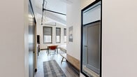 Located on the sixth floor of 305 Michigan Ave in downtown Detroit, this studio unit offers a well-designed, open-plan layout that efficiently combines the living, dining, and kitchen areas into a single cohesive space. Soaring ceilings with exposed industrial-style piping and track lighting lend a loft-like character, while large windows fitted with roller shades invite generous natural light and frame views of the surrounding cityscape.

The kitchen is fully equipped with stainless steel appliances — including a refrigerator, range, microwave, and dishwasher — and features white cabinetry with gold-toned hardware. A compact dining table with two chairs sits at the center, serving as both a dining and workspace. The living and sleeping area is anchored by a king-sized bed beneath a ceiling fan and wall-mounted mini-split unit, complemented by hardwood-style flooring throughout.

The hallway entrance includes a built-in bench and connects to two closets, offering practical storage. The bathroom is clean and modern, with light wood-look flooring, a white vanity with ample cabinet space, and a glass-doored shower enclosure. The overall aesthetic balances industrial and contemporary design elements, creating a functional and stylish urban living space in the heart of Detroit.