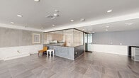 This single-floor commercial space offers a thoughtfully designed, open-plan layout with a distinctly contemporary aesthetic. The space is anchored by a prominent central structure — a glass-enclosed service or display area featuring a sleek dark stone counter, warm pastel tile accents, and full-height glass partitions framed in matte black. This focal element adds both functional utility and architectural interest to the interior.

Large-format gray stone-look tiles extend across the entirety of the floor, lending the space a polished, cohesive appearance. The perimeter walls are finished in a two-tone treatment, combining smooth concrete-style panels on the lower half with a lighter painted surface above, creating a refined yet understated backdrop.

Natural light floods one side of the space through a full wall of floor-to-ceiling windows fitted with roller shades, allowing for flexible light control throughout the day. Recessed ceiling lighting supplements the natural light, ensuring consistent illumination across the open floor plan.

Seating areas are distributed throughout the open space, furnished with mid-century modern-style chairs and occasional accent pieces, suggesting a welcoming, lounge-like atmosphere. Framed artwork adorns the walls, adding warmth and character. The overall design conveys a clean, modern sensibility well-suited for retail, wellness, or professional service use.