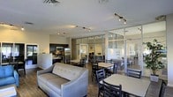 This well-appointed single-floor communal space offers a thoughtfully designed open-plan layout that seamlessly blends relaxation, dining, and productivity. The main area features a comfortable upholstered lounge seating area, ideal for casual gatherings or unwinding, positioned alongside multiple dining tables and chairs that provide flexible seating arrangements for residents or guests. The fully equipped kitchen is outfitted with stainless steel appliances — including a refrigerator, microwave, and range — set against dark cabinetry, making it a functional hub for meal preparation. Track lighting runs throughout the ceiling, providing ample, adjustable illumination across the entire space. A standout architectural feature is the floor-to-ceiling glass partition wall with a sleek metal frame, which separates a private interior room from the main communal area. This enclosed space, visible through the transparent partition, appears to serve as a conference or study room, complete with its own seating and work surfaces. The wood-look flooring ties the space together, adding warmth to the otherwise contemporary aesthetic. Overall, the space strikes a balance between open social areas and dedicated private zones, making it a versatile and inviting environment.