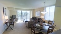This single-floor residence offers a welcoming and well-appointed living environment defined by an open-concept layout that seamlessly integrates the living and dining areas. The living room is anchored by a comfortable seating arrangement and features large floor-to-ceiling sliding glass doors that open onto a private balcony, flooding the interior with generous natural light and providing pleasant views of lush greenery. The neutral-toned walls and plush carpeting contribute to a warm, inviting ambiance throughout the space. A flat-screen television and media console are thoughtfully positioned within the living area, making it both functional and relaxing. Adjacent to the living room, the dining area is centered around a classic round dining table with seating for multiple guests, illuminated by an elegant pendant light fixture that adds a refined touch to the space. A framed piece of artwork on the wall ties the two areas together visually, enhancing the cohesive design. Additional windows along the side wall further brighten the interior, ensuring the space feels open and airy. Overall, this residence combines practical functionality with a comfortable, light-filled atmosphere well-suited for everyday living and entertaining.